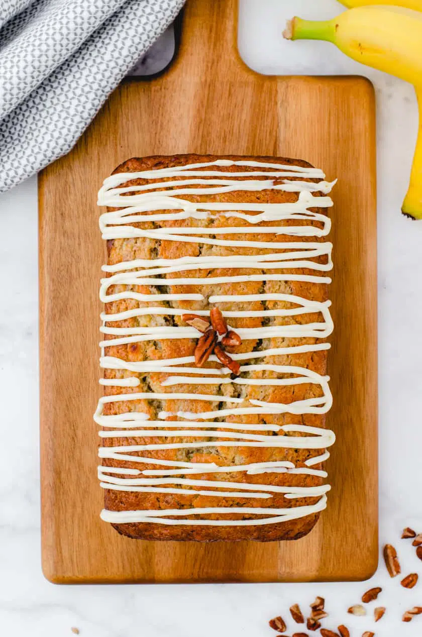 Buttermilk banana bread on a wooden cutting board with cream cheese icing drizzled over it and walnuts decorating the top.