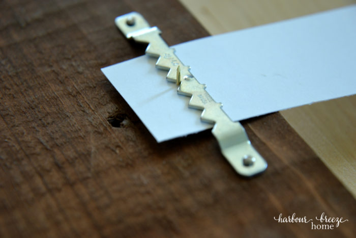 How to hang pictures on the wall: Are you paralyzed from hanging pictures on the wall for fear you'll put a nail in the wrong place? If so, this life hack will rock your socks off! Learn how to quickly and simply hang pictures in exactly the place you want using simple tools around the house. Click to read the easiest method ever! Close up of the thumbtack and paper under the sawtooth hanger (how to hang a picture on the wall)