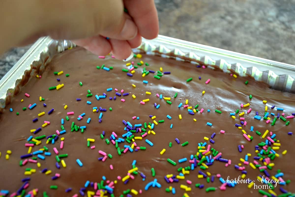 Multi-colored sprinkles being sprinkled on a pan of chocolate brownies with icing. Texas brownies makes a great Summer dessert for a party!