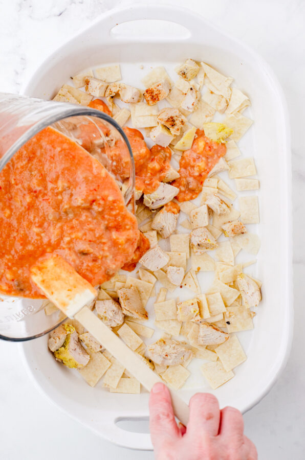 A woman pouring sauce over the layers of Mexican chicken casserole in a baking dish.