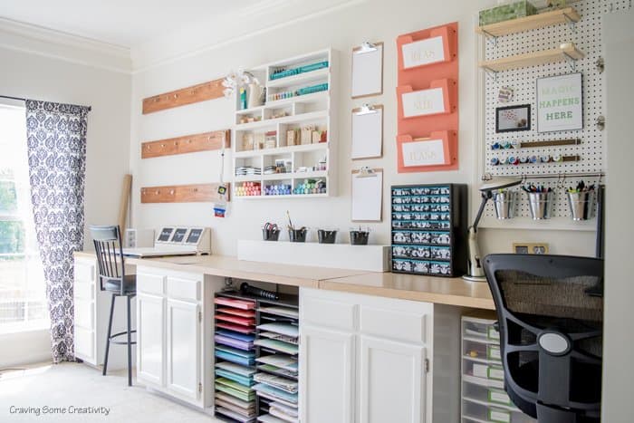 Craft supplies like scrapbooking paper and accessories organized against one long wall with cabinets below and hanging pegboard and shelves above