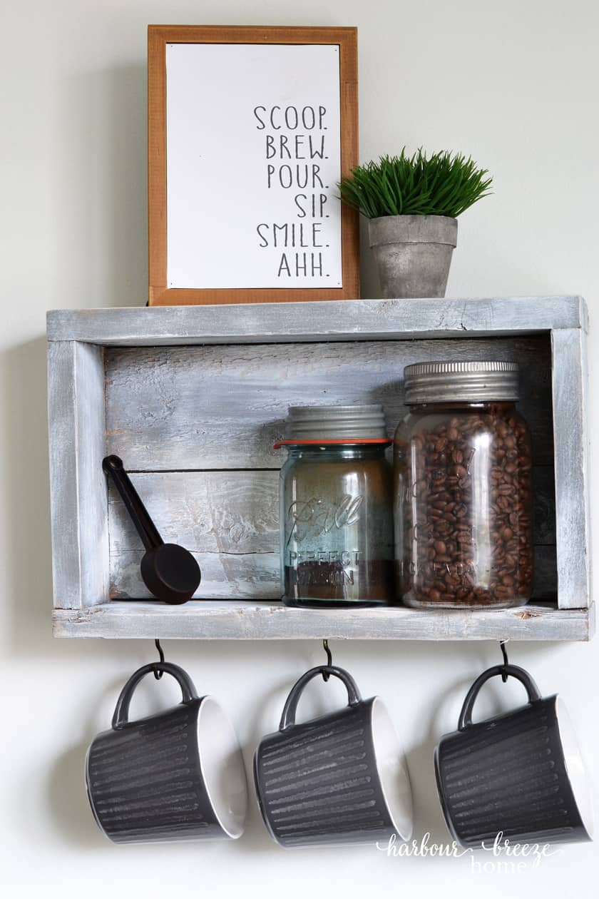 Rustic wall shelf with 3 hooks holding coffee cups and on the shelf is sitting antique mason jars with coffee beans in it.