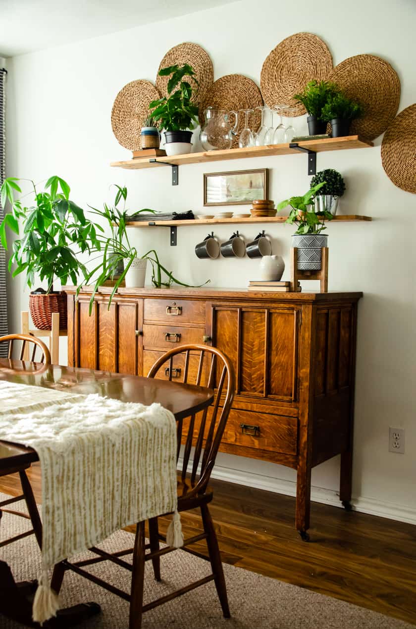 boho dining room decor featuring an antique wooden buffet, farmhouse shelves, vintage art, woven placemats on wall, real and faux plants and glassware