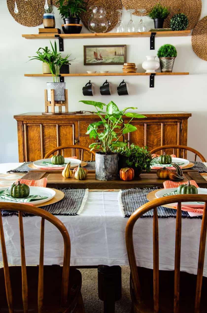 Dining room table set in a Fall decor scheme with faux pumpkins, live plants, orange checked napkins, and floral green napkins.