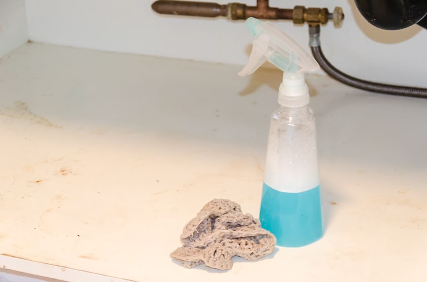 Dirty under kitchen sink cabinet with a cloth and bottle of cleaning spray beside it.