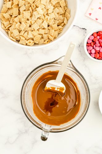 Melted chocolate mixture in a microwave safe bowl with a large bowl of cereal mix behind