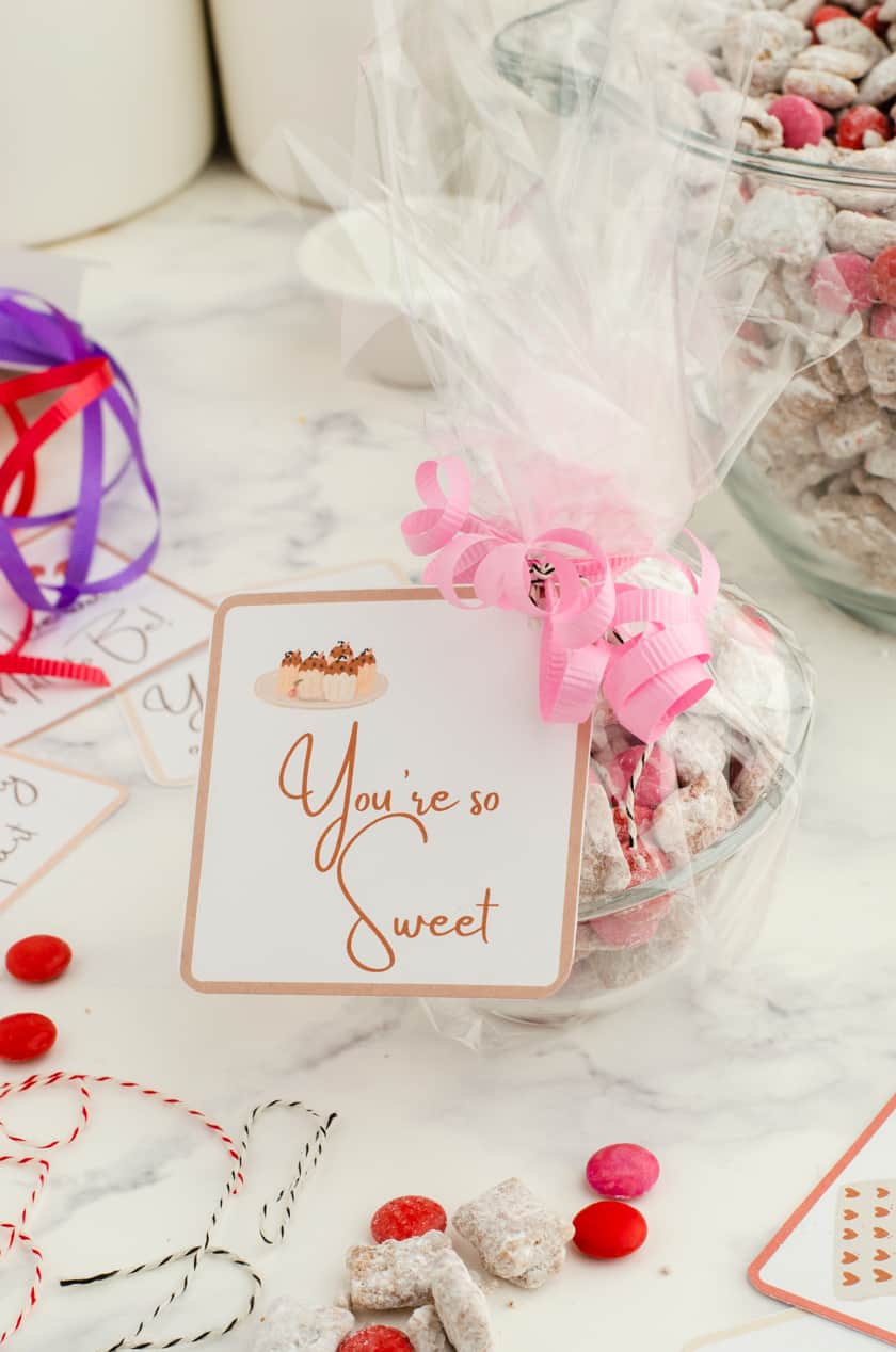 a clear bowl filled with valentine puppy chow snack mix with cellophane tied around it with a pink ribbon and valentine card