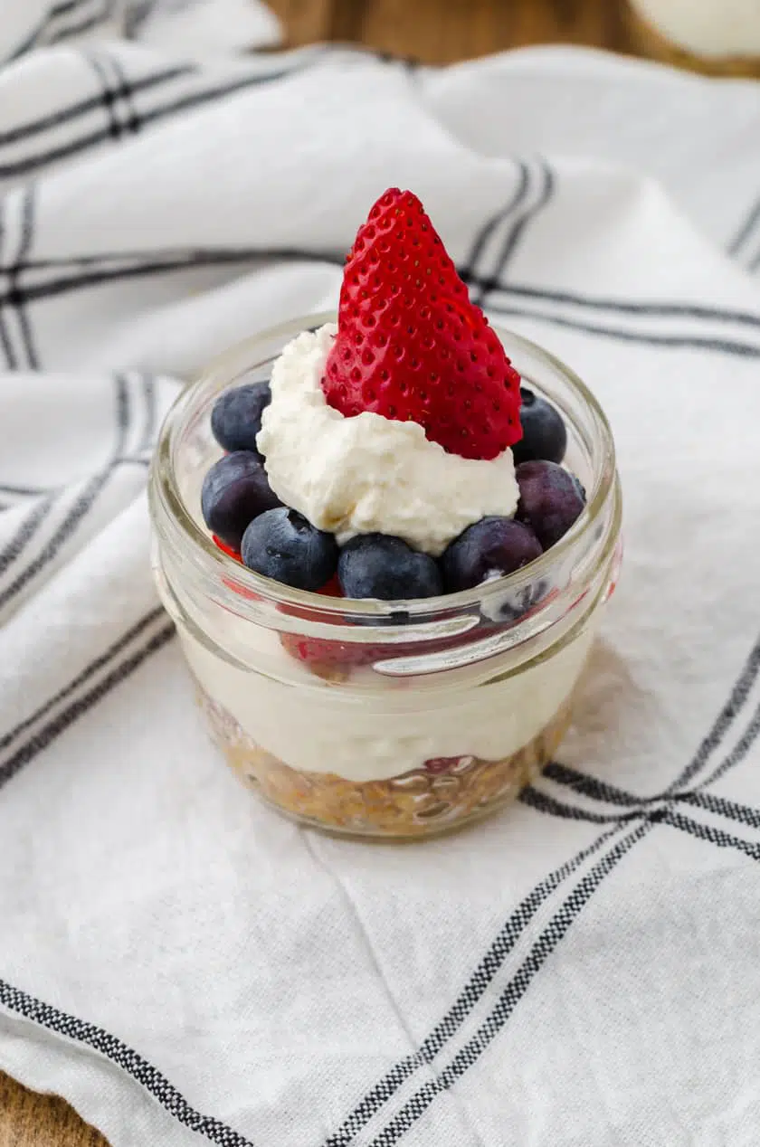 No bake cheesecake in mini mason jar with a strawberry on the top surrounded by blueberries. It's a red white and blue dessert!