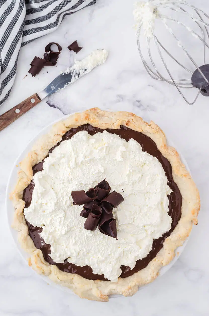 A whole chocolate pudding cream pie with a mixing bowl whisk with whipping cream beside.