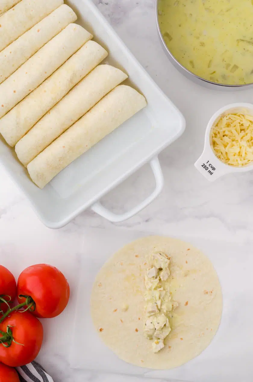 Creamy Chicken enchiladas rolled up in a baking pan with a pot with the creamy sauce beside.