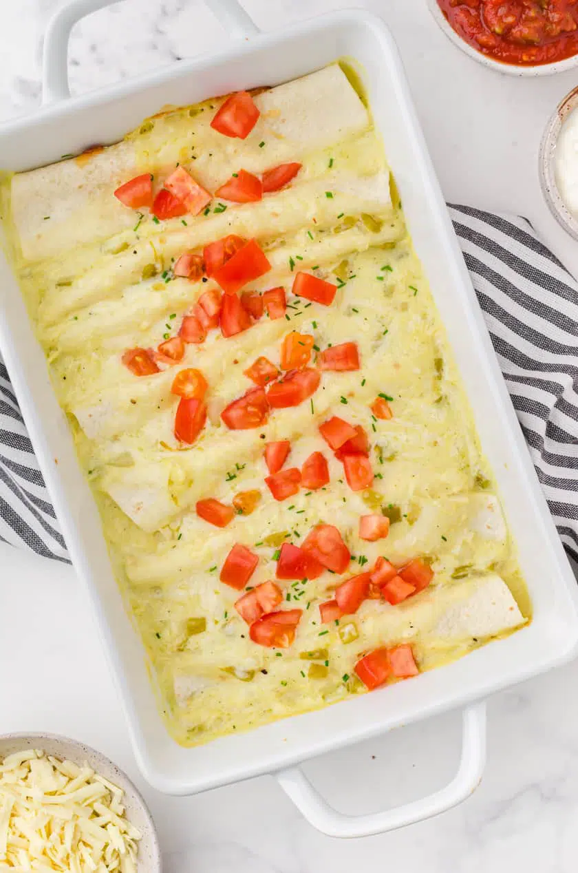 A pan of baked creamy chicken enchiladas with a garnish of chopped tomatoes and cut up chives on the top.