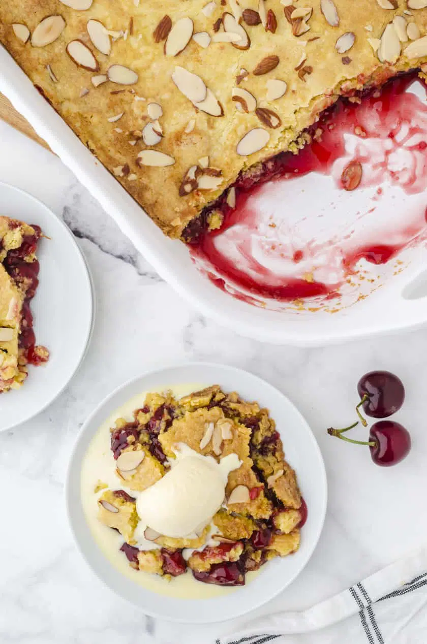 a pan of cherry dump cake with a few servings taken out and a slice of cherry dump cake on a dessert platter with vanilla ice cream melting over the top of it.