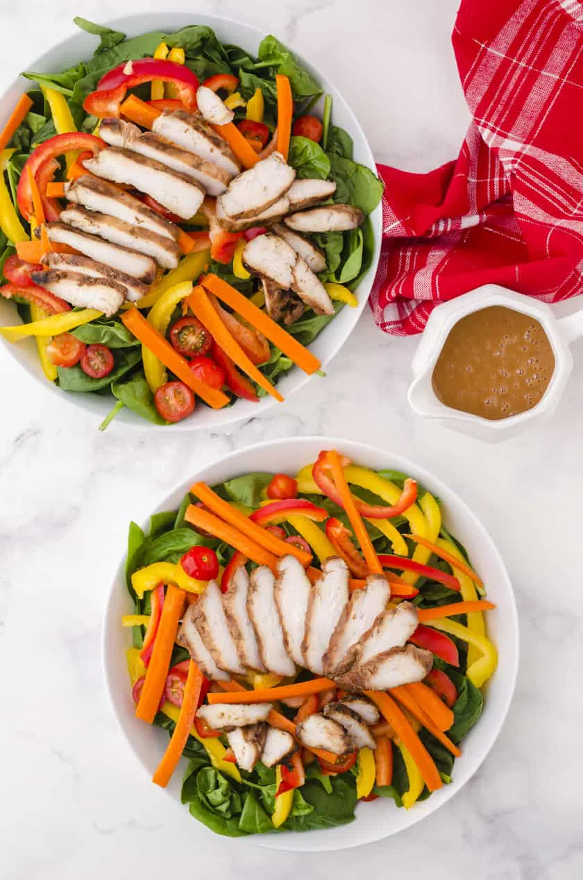 Two plates of grilled chicken salad with the pitcher or peanut chili sauce sitting beside them