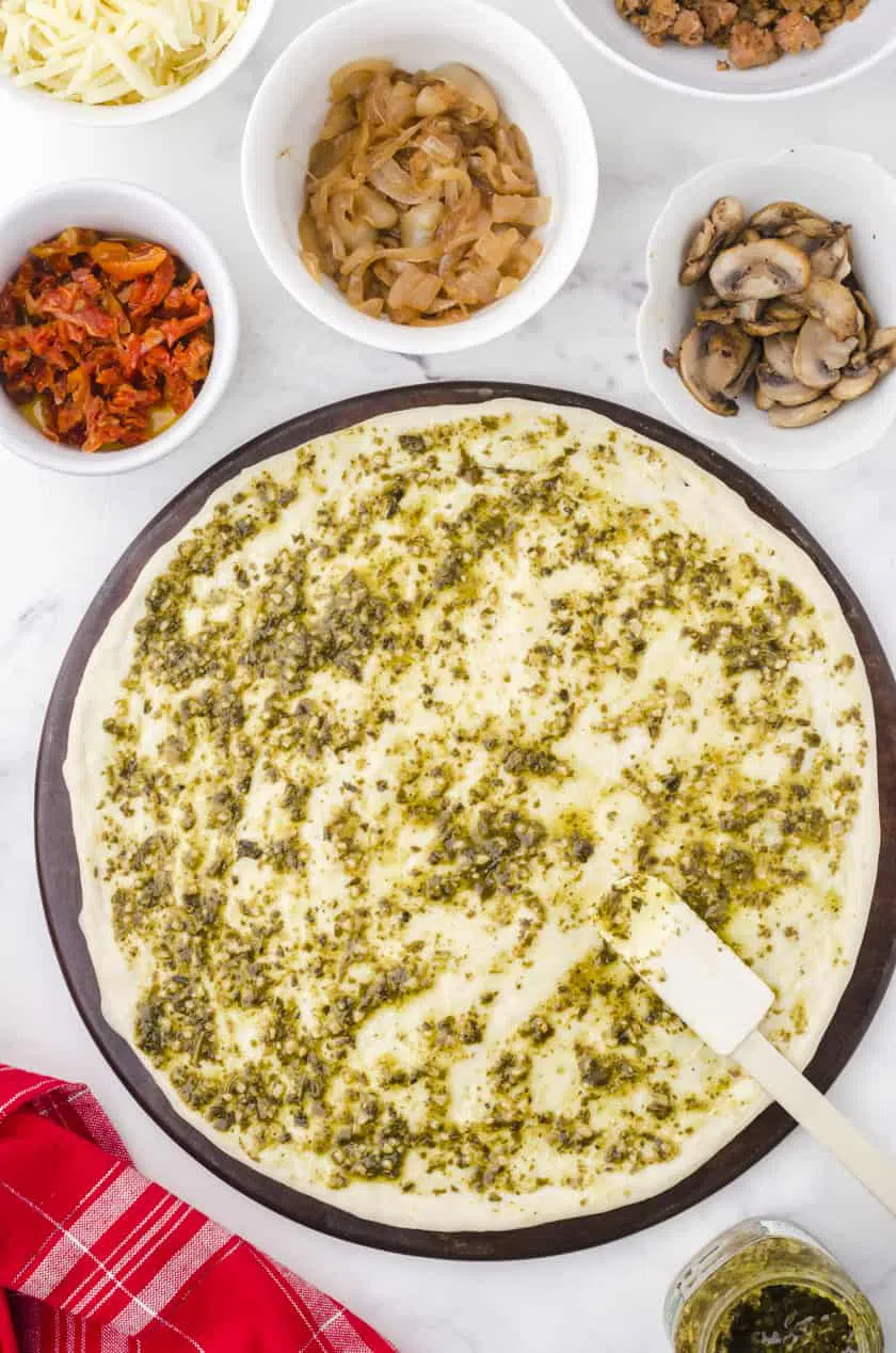 Pesto sauce being spread on a base of homemade pizza dough with dishes of toppings around it.
