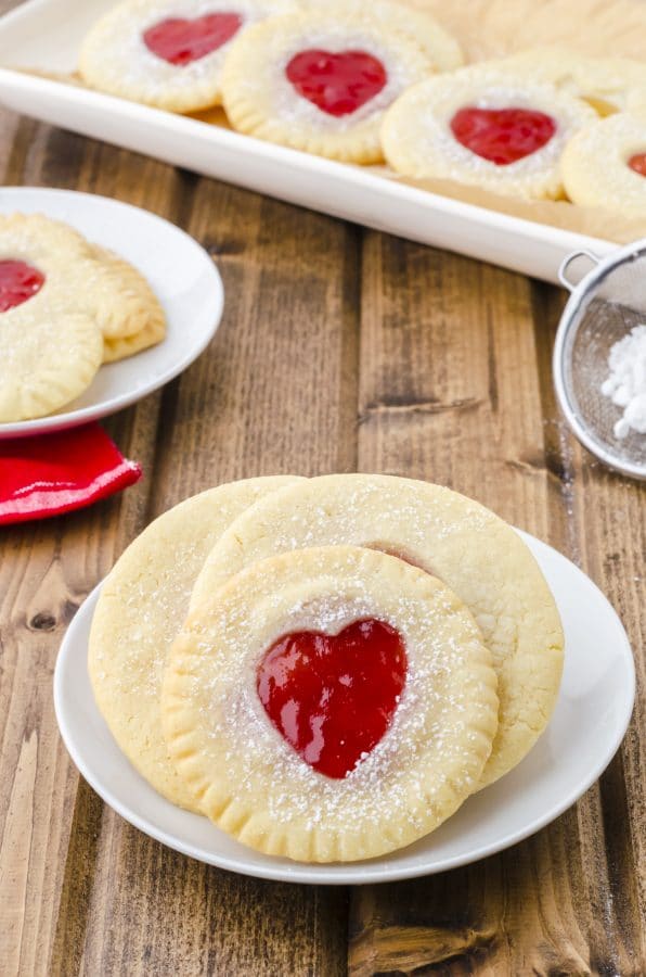A plate full of rounc jam filled sandwich cookies with strawberry jam filling a heart shaped cutout.