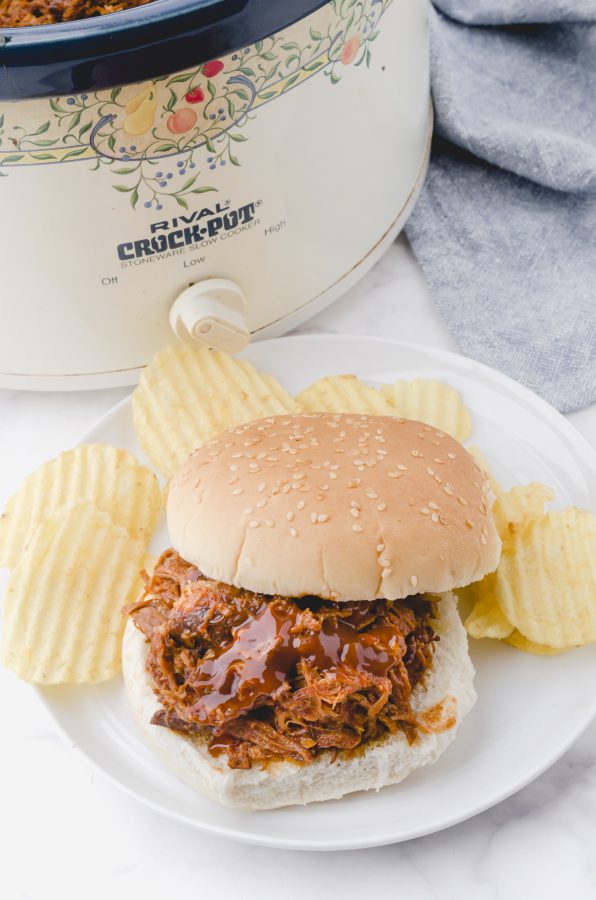 Pulled pork piled on a bun to make a pulled pork sandwich with potato chips on the side of the plate.