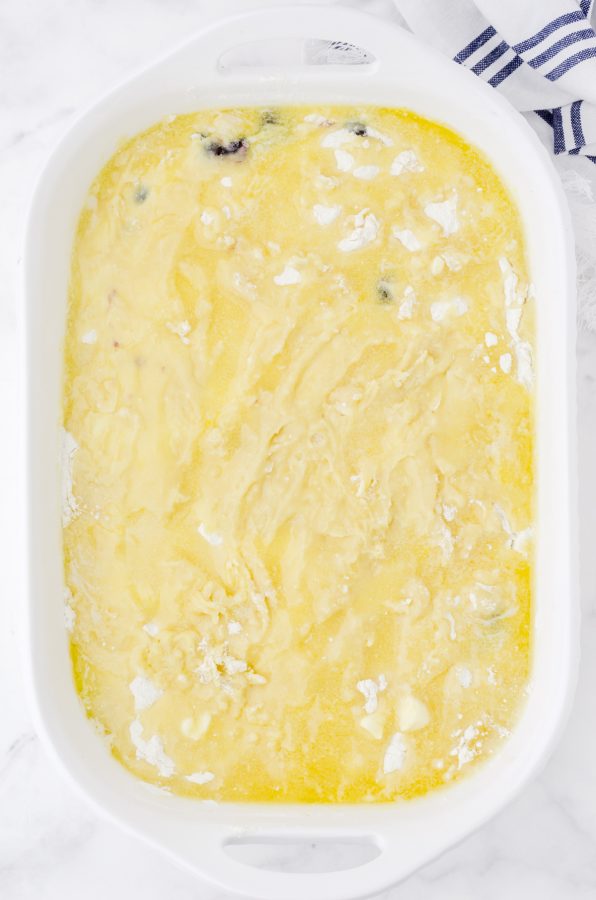 A white 9 x 13 baking dish with blueberry dump cake ready to be put in the oven. The butter is melted and drizzled over the top to almost completely cover the cake mix powder.