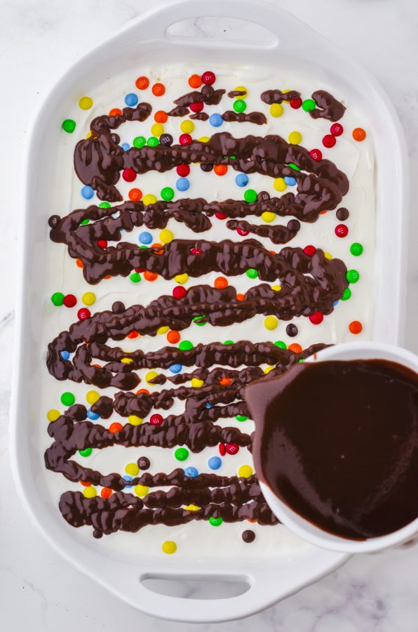 hot fudge sauce being drizzled over the m&ms layer of an oreo ice cream cake