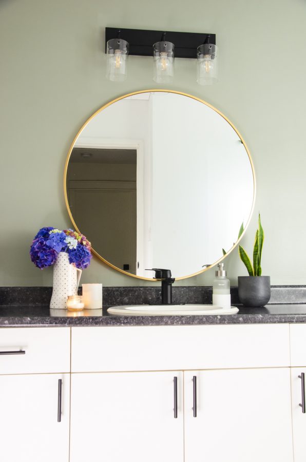 The vanity area with a round mirror with brass frame and new light fixtures, and the wall painted in evergreen fog by Sherwin Williams.