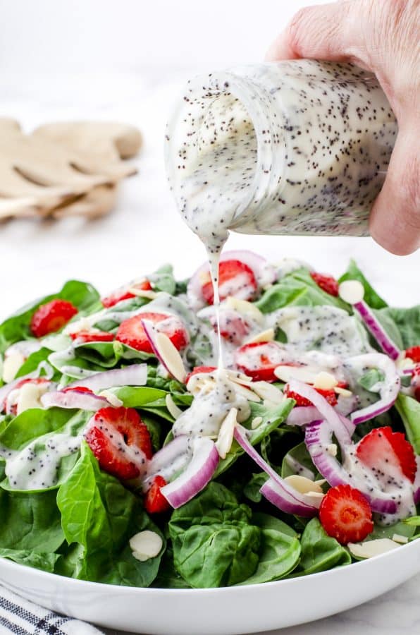 Creamy poppy seed dressing being drizzled onto a spinach and strawberry salad.