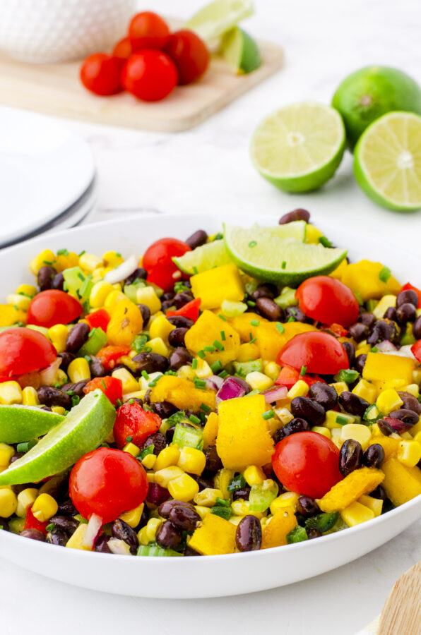 A bowl of mango salad with black beans garnished with lime slices, cherry tomatoes, and fresh chives.