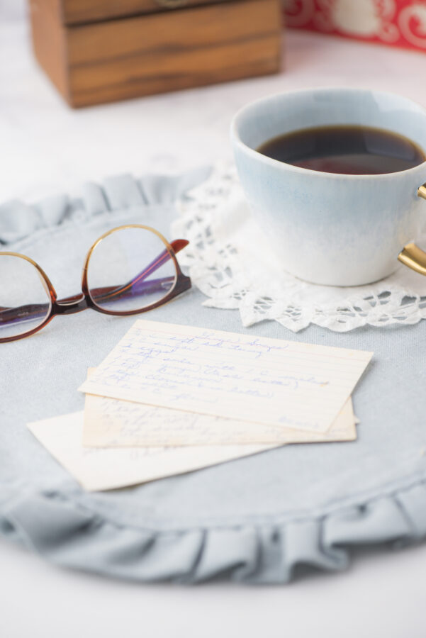 a pile of old recipe cards with a cup of coffee and pair of glasses beside it. Vintage cookbooks are in the background.
