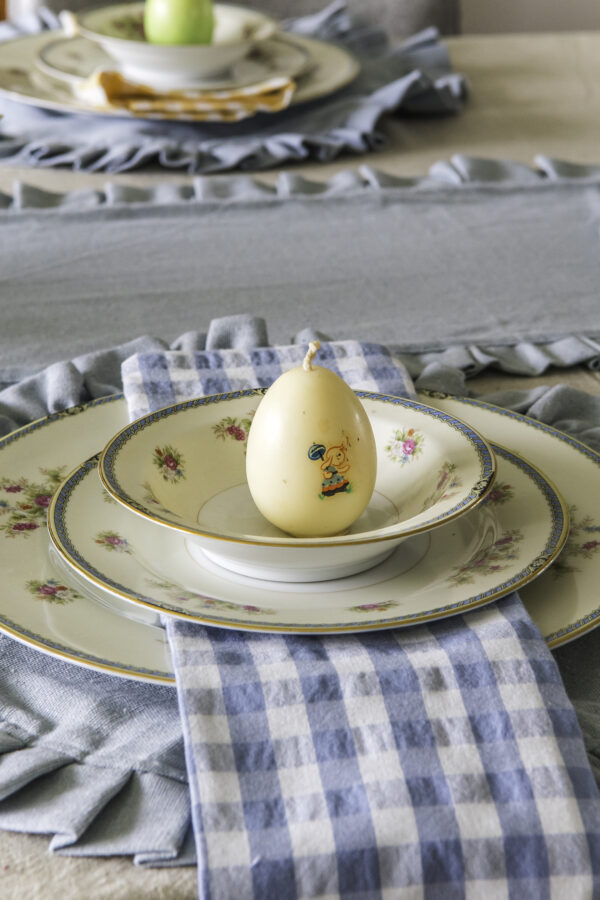 A table setting idea with a blue round ruffled placemat, blue and rose vintage china, blue checked cloth napkin, and a vintage egg candle sitting on the top of the top plate.