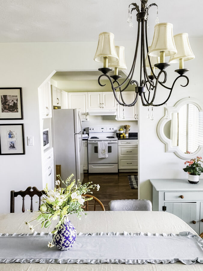 Looking into the doorway to the kitchen from a dining room - showing the table with an arrangment on it and a mirror hanging above a small dresser on one side.