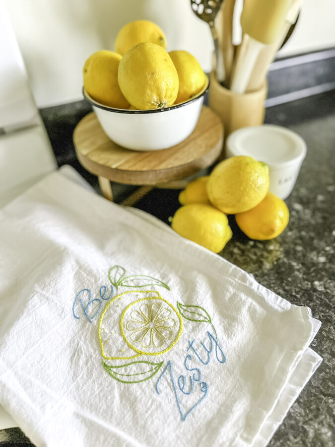 A bowl of lemons with a tea towel beside that says, "be zesty" on it.