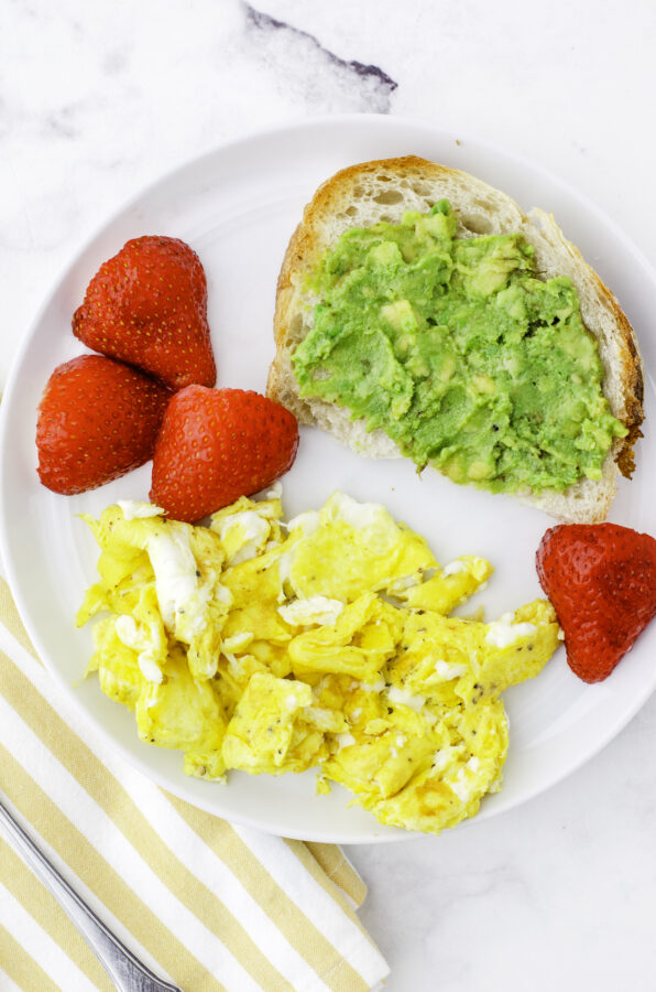 Mashed avocados on a piece of sourdough toast on a plate with scrambled eggs and strawberries.