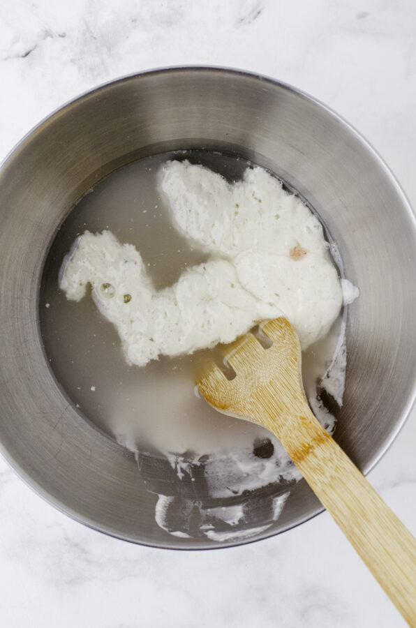 Water, salt, and sourdough starter being mixed in the mixing bowl of a stand mixer with a wooden spoon with holes in it. Step 1 of mixing sourdough bread dough.
