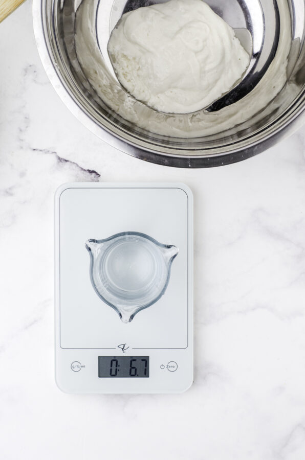 A container of water being measured on a scale with a bowl of sourdough starter behind it. Step 2 of feeding sourdough starter.
