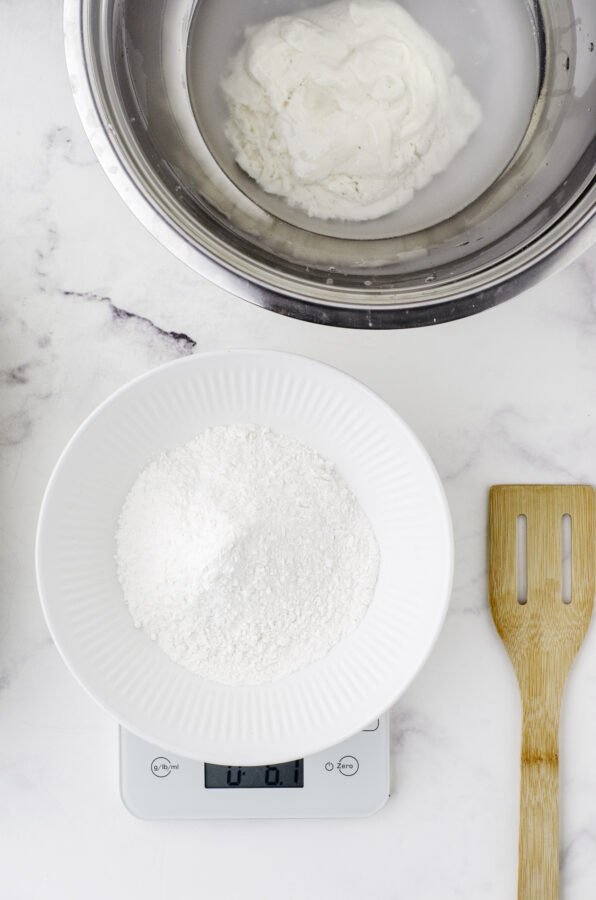 A bowl of flour being measured on a kitchen scale with a wooden spoon with holes beside it. A metal bowl with sourdough starter and water is behind it. Step 3 of feeding sourdough starter.