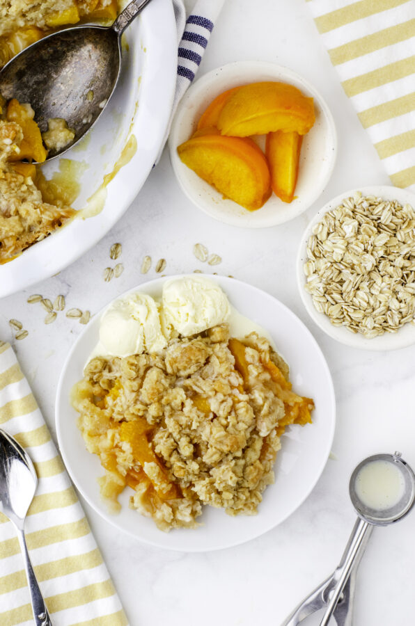 A serving of peach crisp with scoops of ice cream beside it on a dessert plate, with a pan of peach crisp with a scoop out of it above the dessert plate.