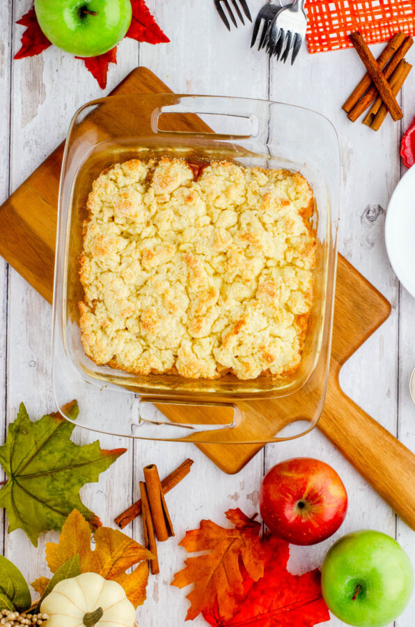 A pan of apple cobbler on a table surrounded by plates, cinnamon sticks, forks, napkins, and fall leaves.