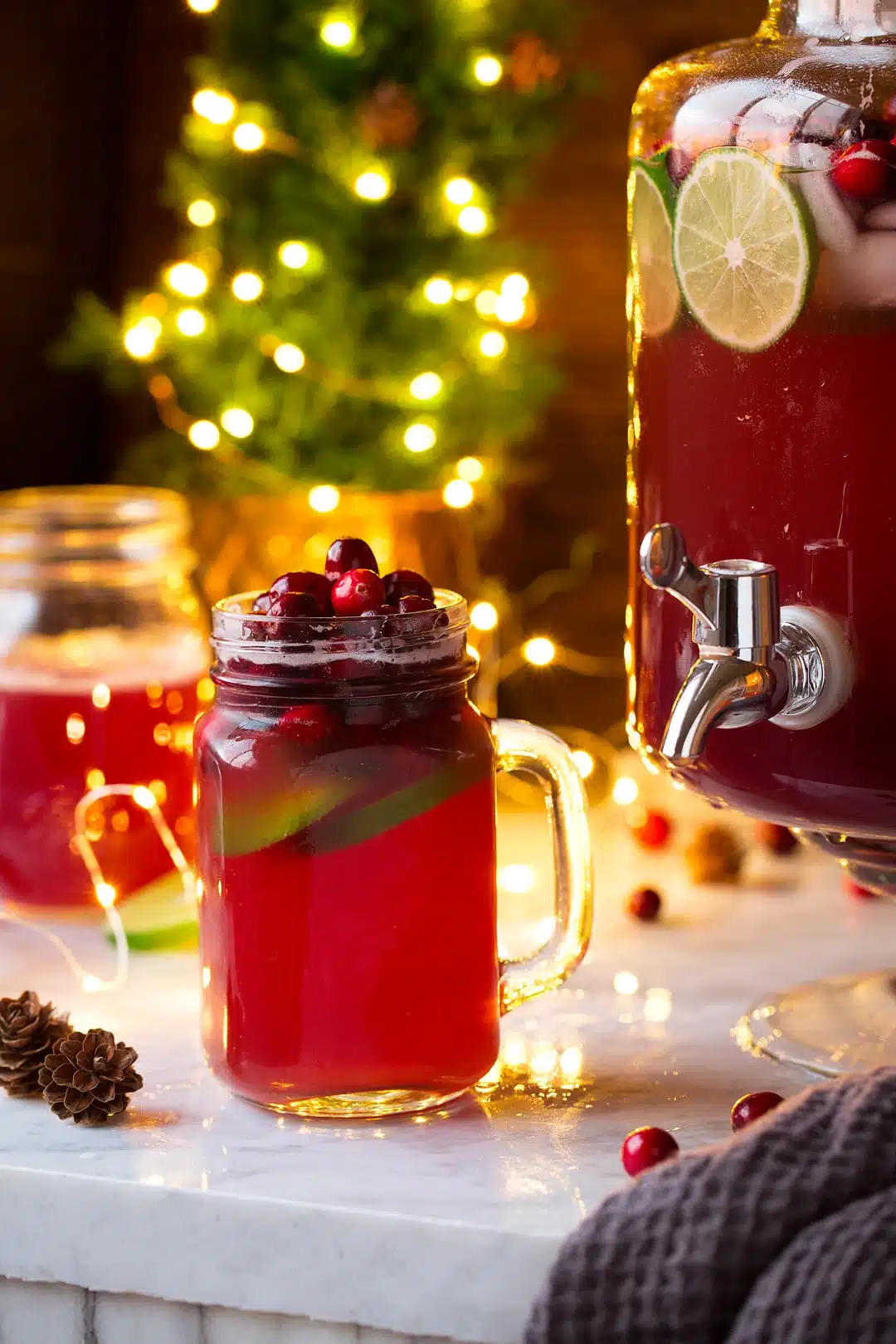 Glass of Christmas punch beside a large dispenser with punch in it.