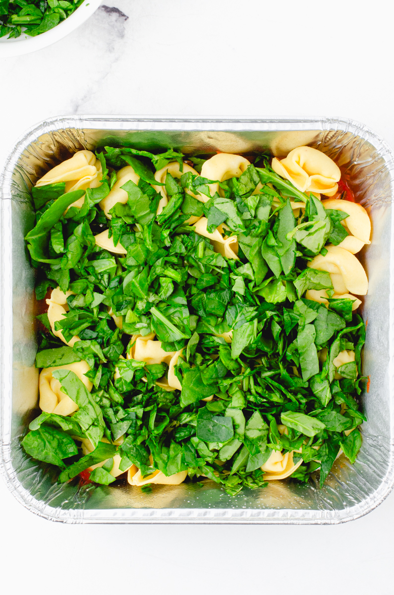 Chopped spinach on top of tortellini in a square pan for tortellini casserole