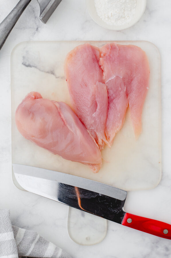 Chicken breasts on cutting board being butterflied with a large knife.