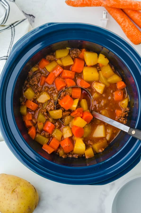 a crock pot full of cooked hamburger stew with some carrots and potatoes sitting beside it.