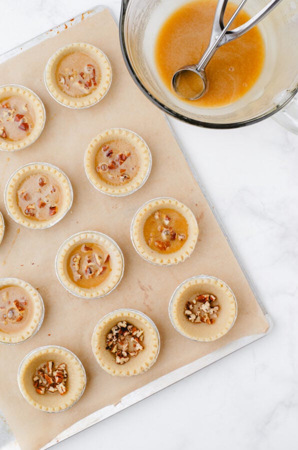 Butter tart shells with chopped pecans in the bottom with a bowl of butter tart filling beside with a cookie scoop in it.